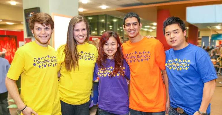 Photo of five young adult volunteers standing together in the library and wearing bright tshirts that say Reading Rocks and 