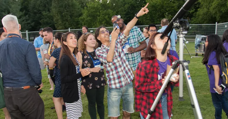 Photo of a group of people standing outside and pointing up into the sky.  In the front, a woman looks through a telescope.