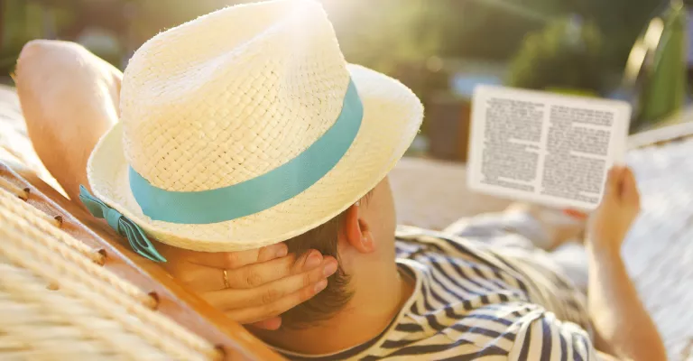 Person lying in a hammock and reading an ebook. They are wearing a straw hat and the sun is shining brightly.