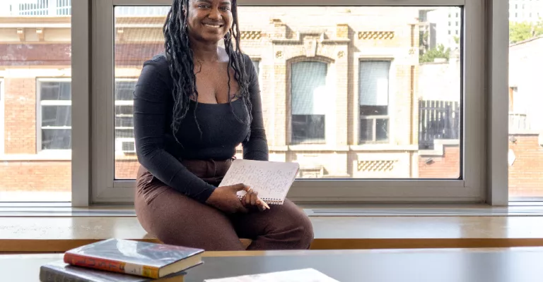 Woman sitting in front of window with streetscape behind her, hodling a notebook.