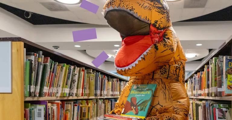 Large cartoonish dinosaur suit standing among library shelves holding children's books.