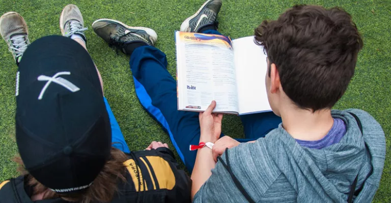 Two boys sitting outside on grass reading a book