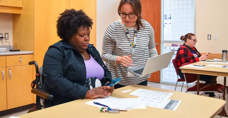 Woman in wheelchair at a table holding a phone with another woman standing next to her holding a laptop