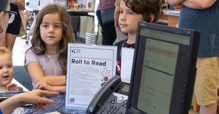Two kids standing at Library desk registering for the Summer Reading Club