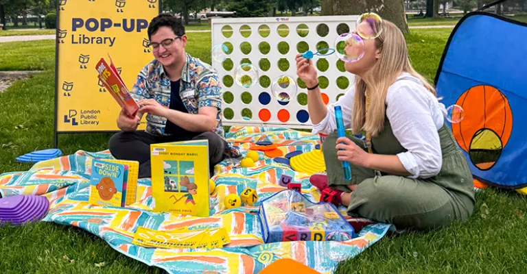 Library staff sitting on a blanket at a park surrounded by large games, books and activities. One is reading a book and the other is blowing bubbles.