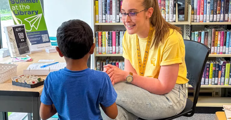 Library staff member sitting in a chairs across from a young child, registering for the Summer Reading Club. 
