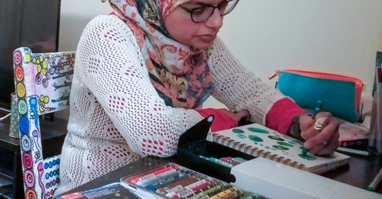 Young Muslim woman sitting in a chair at a table with art supplies in front of her, colouring in a sketchbook. 