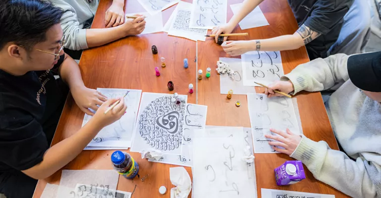 Group of people around a table using qalams or reed pens to create Islamic calligraphy