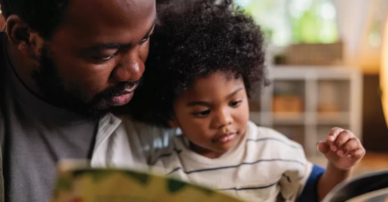 A young child sits on his father's lap and reads a picture book together.
