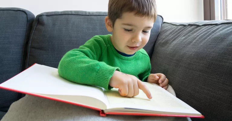 A boy about 6 or 7 years old reads a book using his finger to follow the text. 