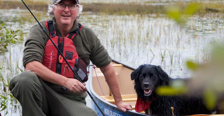An older man is holding a fishing rod while kneeling at the edge of a pond. Also pictured is a blue canoe with a dog sitting in it. 