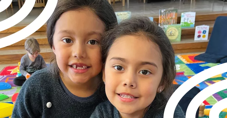 Two young children smile at the camera during a Library program, standing on a colourful play mat with books and toys visible in the background.