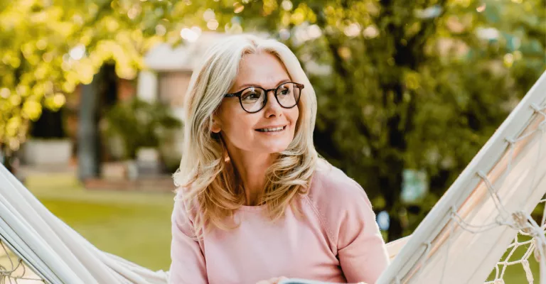 Smiling senior blond woman wearing glasses while reading in hammock in the summer garden