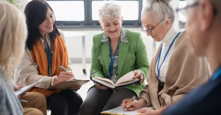 A group of older adults sit together in a bright room, reading and writing in notebooks during a discussion. Two people in the centre look down at an open book.