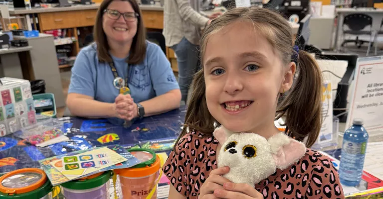 A smiling child holds a small white stuffed animal at a Library service desk. Behind her, a staff member sits beside Summer Reading Club materials, prize containers, and activity supplies.