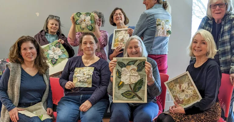 Group of smiling adults seated and standing in a program room, holding up textile art pieces with floral designs. One participant wears a denim jacket with a fabric flower design on the back.