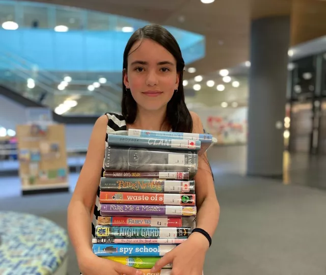 Young person on the main floor of Central Library holding a very large stack of books and smiling.
