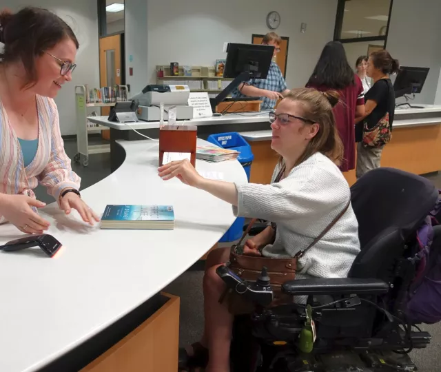 Person using wheelchair checking out book from Central Library with Library staff person at checkout counter