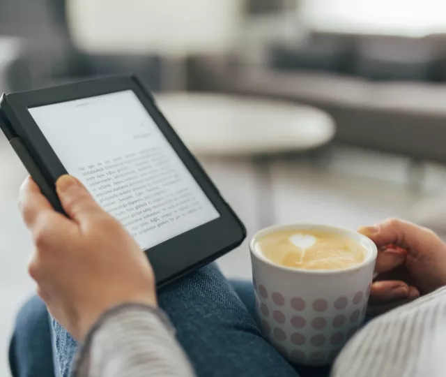 A photo of someone's hand holding a tablet with an ebook displayed. In the other hand they hold a cup of coffee which sits on their lap.