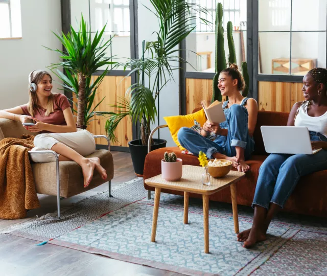 A photo of three young women sitting in a living room. One is holding a cellphone one and wearing headphones one is holding a laptop and one is holding a book. They are smiling and looking at each other.