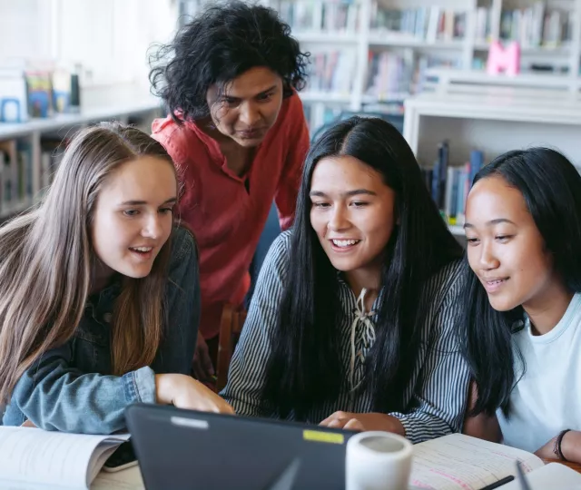 Four older students sit at table looking at laptop and smile.  In the background are bookshelves.
