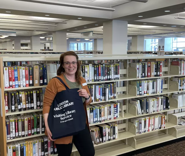 Photo of a librarian holding a Visiting Library Service black bag and standing in front of a book shelf at the Library.