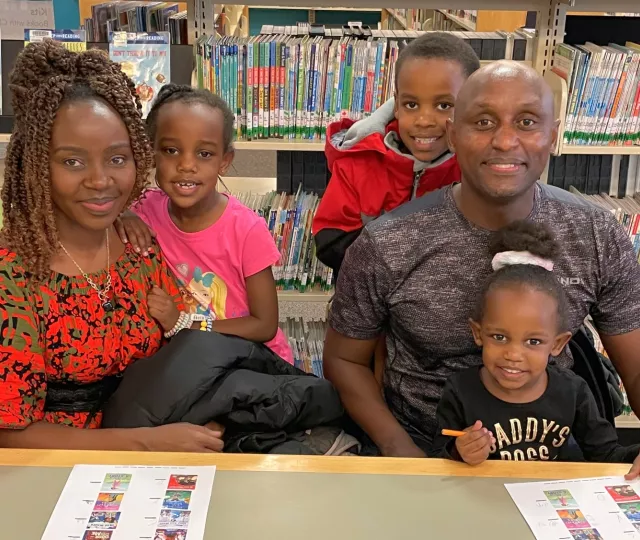 Photo of a family of two adults and three young children sitting at a table in the Children's Library. In the background are bookshelves.