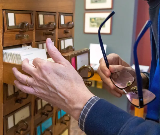Photo of a librarian's hand flipping through index cards in large wooden card catalogue in the London Room. In the other hand, he holds his glasses.