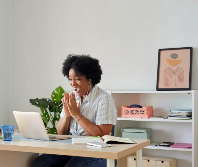 Photo of a woman looking at her laptop and smiling and clapping. In the background are bookshelves and on the table are books, a phone and a glass.