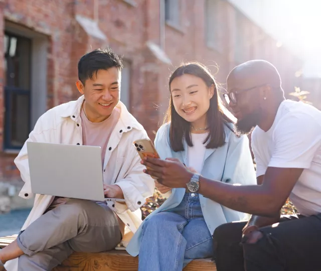 Photo of three people sitting outside in sunshine. One person holds a laptop, another a phone and the third, a tablet.