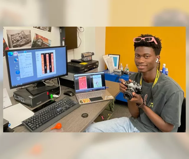 Photo of a young man holding a camera and sitting at a table with a computer and laptop. In the background is more digital equipment.