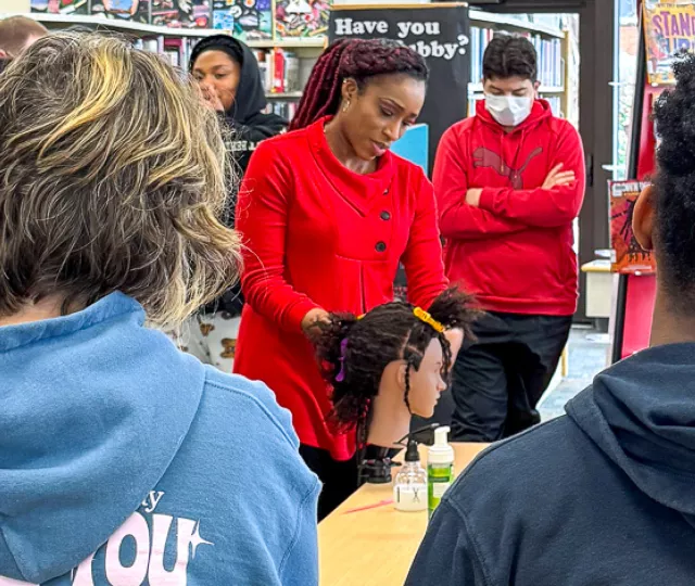 Students gather around Lana as she braids a mannequin's hair in the Library.