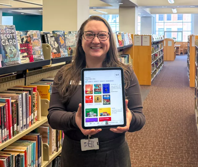 Librarian standing in between book shelves holding out an iPad with the Libby app loaded.