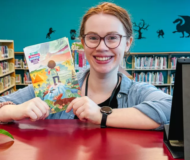 Claire holds up a TD summer flyer in the Children's area, surrounded by bright colourful books.