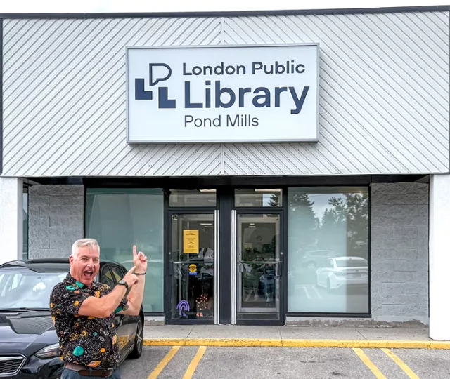 Kevin Moore stands in front of Pond Mills Branch Library, pointing at the front doors.