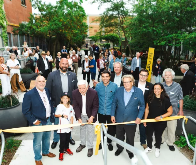 Local dignitaries donors and Library staff gather behind a yellow ribbon as a young boy holding scissors prepares to cut the ribbon in the Rotary Reading Garden.