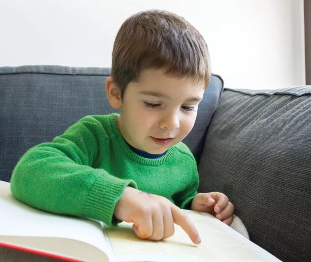 A boy about 6 or 7 years old reads a book using his finger to follow the text. 