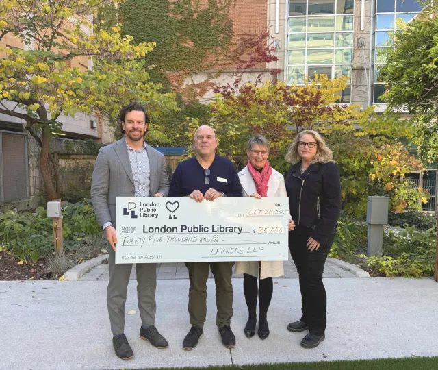 A representative from Lerners LLP stands with three representatives from London Public Library in the Rotary Reading Garden holding a large cheque for twenty five thousand dollars.