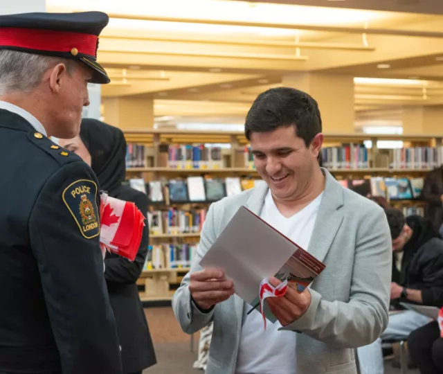A man holds his citizenship certificate and smiles while a London Police Services Officer in uniform looks on. In the background participants sit on chairs surrounded by bookshelves.