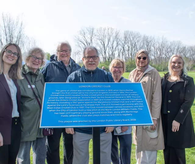 7 people, members of the Historic Sites Committee stand behind a blue plaque. In the background is parkland.