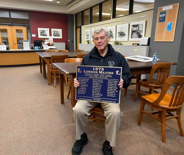 Barry Boughner sits in the London Room at Central Library holding a blue commemorative plaque recognizing the 1975 London Majors championship team. Research tables, archival storage cabinets, and historic photographs line the room behind him.