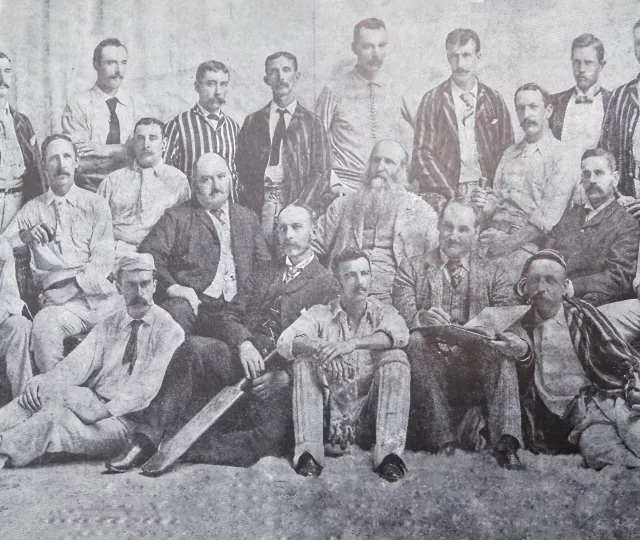 Balck and white undated photo of Cricket players in white uniforms and caps, some wearing striped balzers and some in full suites.
