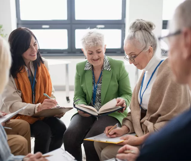 A group of older adults sit together in a bright room, reading and writing in notebooks during a discussion. Two people in the centre look down at an open book.