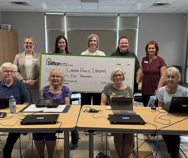 3 members of the Sifton Family Foundation Board along with a facilitator and a London Public Library staff member stand with a large cheque. In front of them, 4 senior students sit with laptops in front of them.