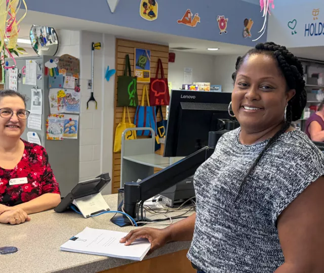 Library staff member stands behind a counter and smiles next to featured patron Shelly at Masonville Branch Library