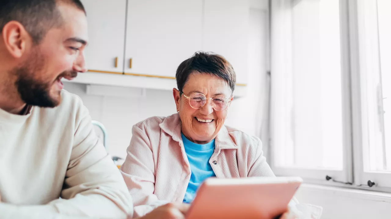 Older adult with table sits at table in kitchent showing tablet to adult