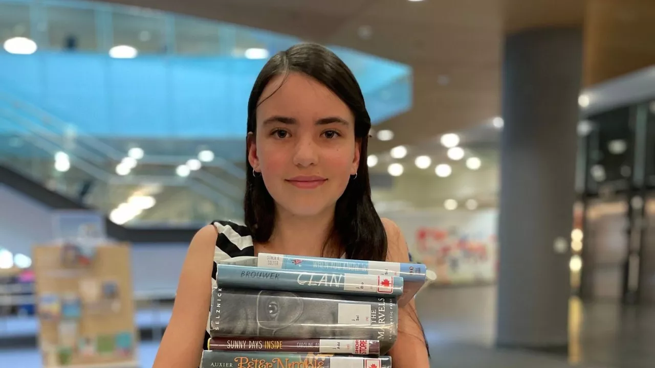 Young person on the main floor of Central Library holding a very large stack of books and smiling.