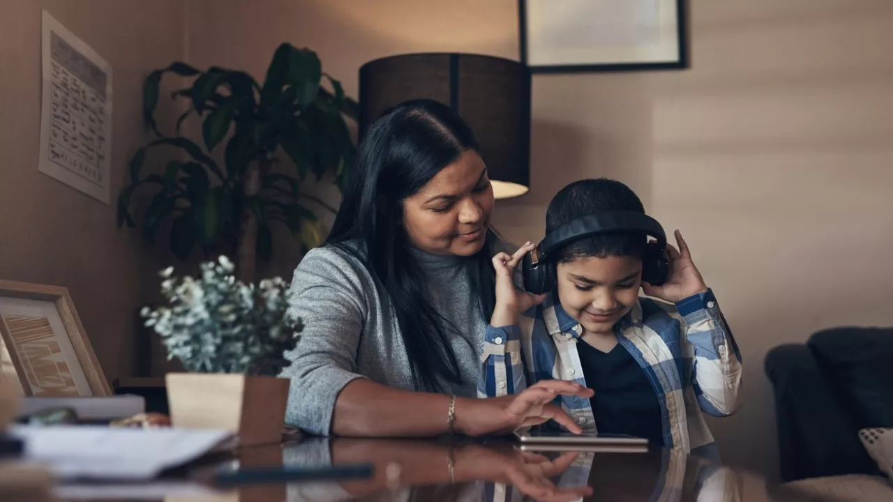 Photo of a mother and child using a tablet on a table. The child is wearing headphones.