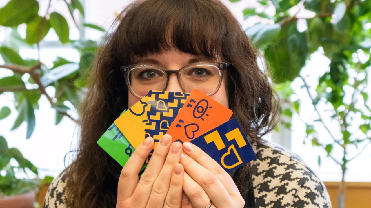 Photo of a woman wearing glasses and holding 5 differently designed London public Library cards in front of her face. In the background are plants and a window.