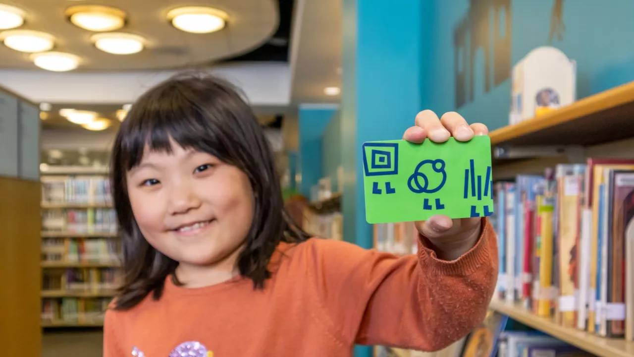 Photo of a child holding a green London Public Library card in their outstretched hand. In the background are bookshelves.